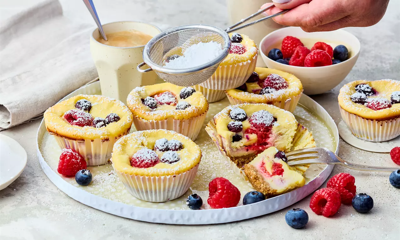 Puderzucker wird mit Sieb aufgestreut auf Mini-Cheesecakes mit Keksboden, cremiger Frischkäse-Quark-Füllung und frischen Beeren wie Himbeeren und Blaubeeren – einfach im Muffinblech backen.
