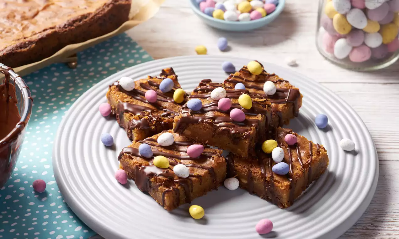 Golden Easter blondie squares drizzled with dark chocolate, topped with pastel-coloured candy eggs, arranged on a white plate