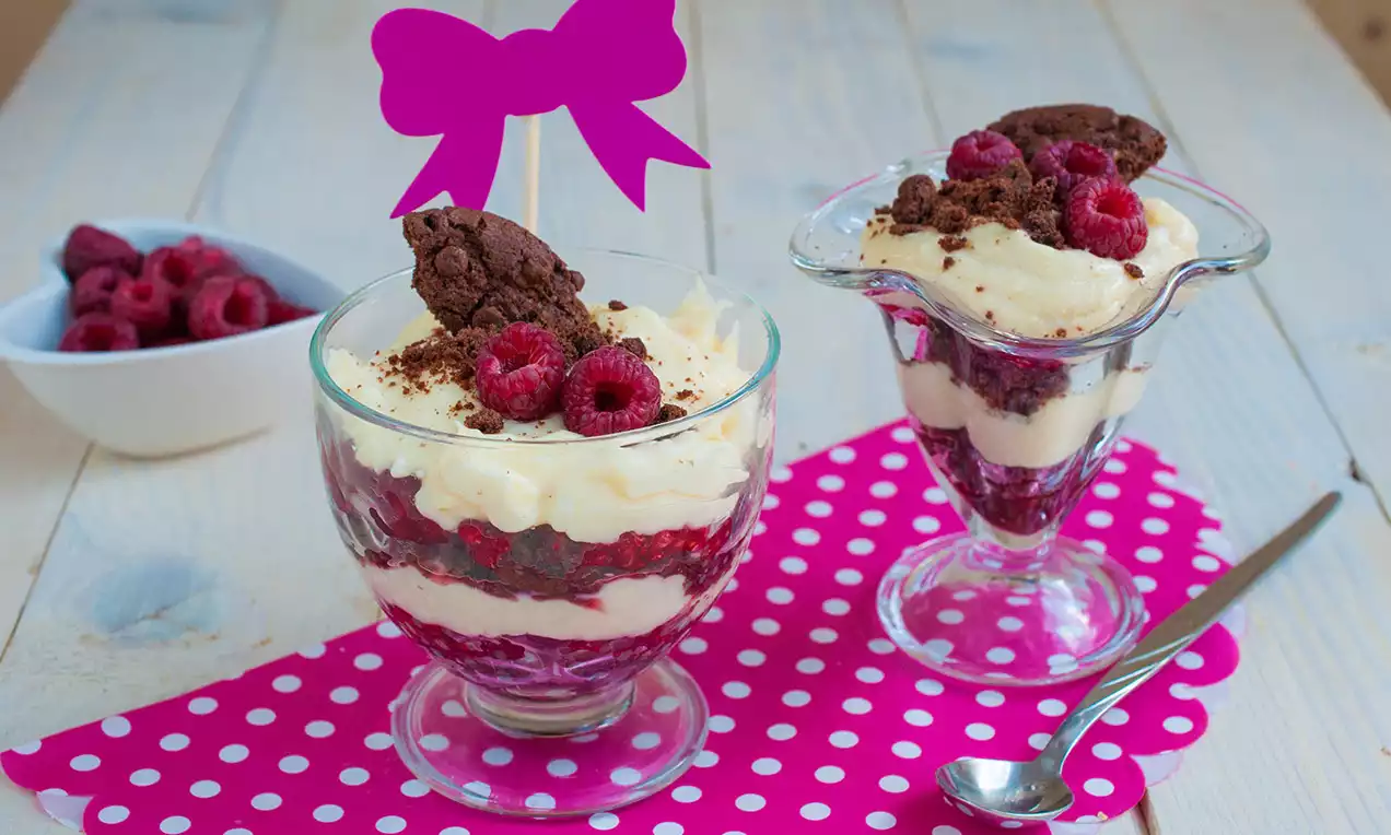 Sklenice svrstvené pěny světlé barvy, malinové vrstvy, čokoládových drobků, ozdobené malinami a sušenkami na růžovém puntíkatém ubrousku