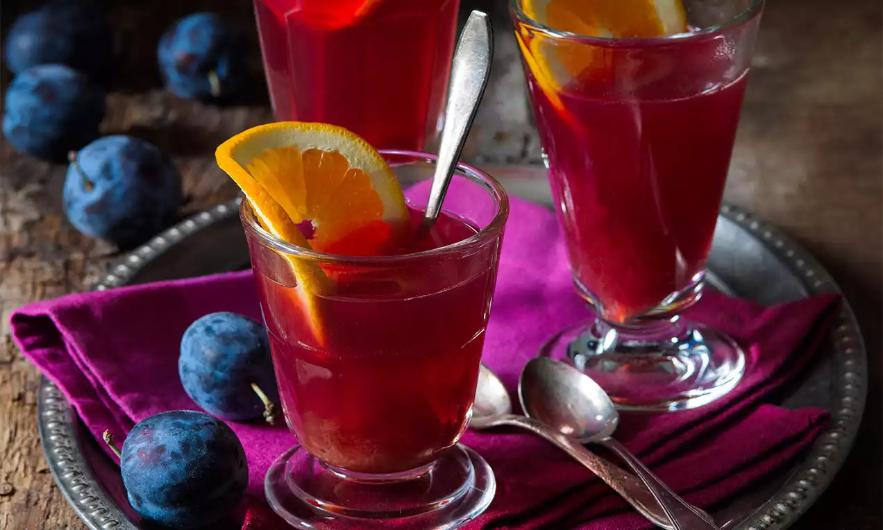 Teplý tmavorubínový nápoj s plátkami pomaranča v sklenených pohároch, na fuchsiovom obrúsku s čerstvými slivkami a lyžičkami na tácke