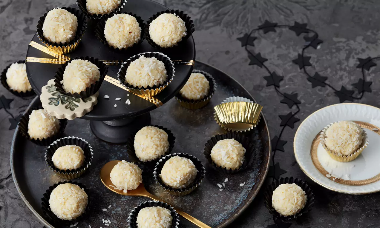 Kugelförmige Eierlikör-Trüffel mit weißer Kokosraspel-Textur, in schwarzen und goldenen Papierförmchen auf Tellern und Etagere arrangiert