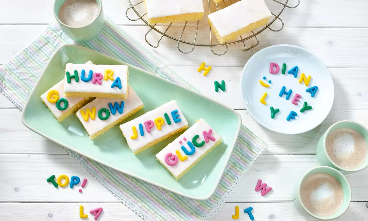 ABC-Kuchen mit weißer Glasur und bunter Buchstaben-Dekoration auf einem hellgrünen Teller, umgeben von Tassen und gestreiftem Tuch