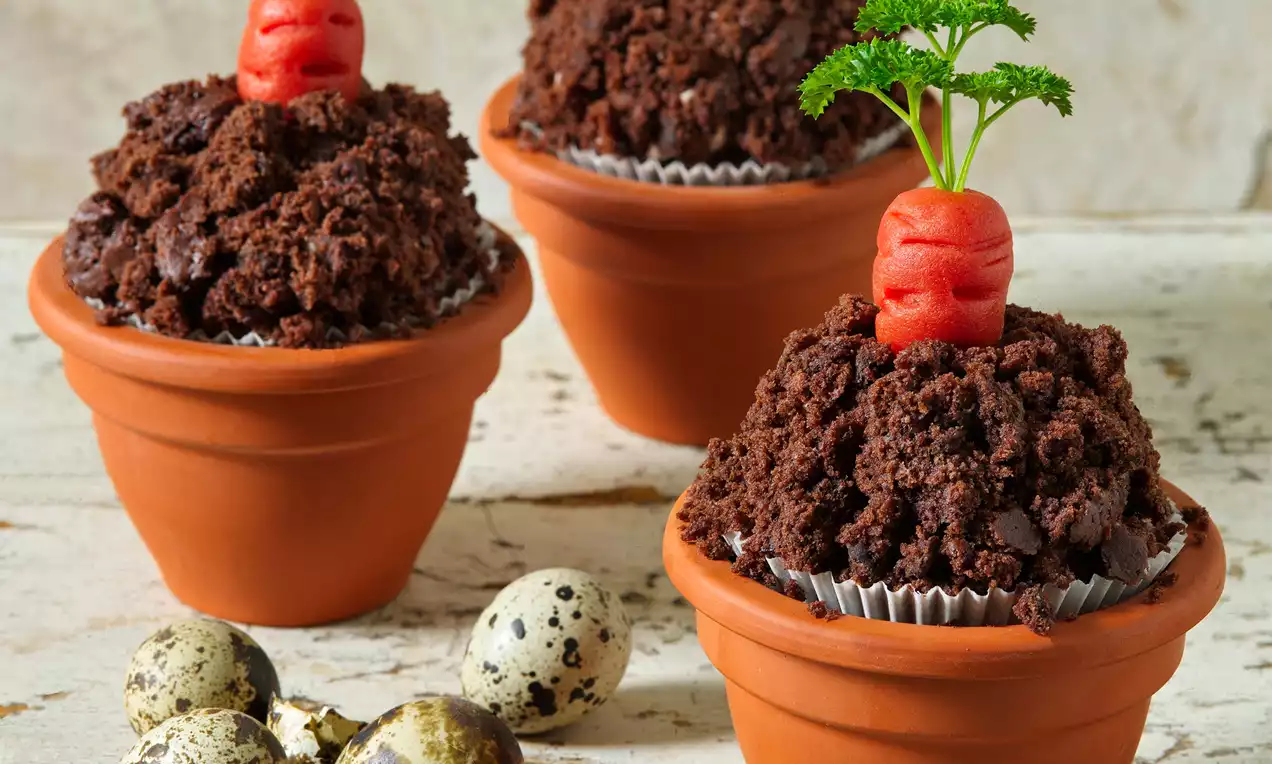 Čokoládové muffiny s tmavohnedou drobivou textúrou, servírované v terakotových kvetináčoch, s marcipánovou oranžovou mrkvou a zelenou vňaťou
