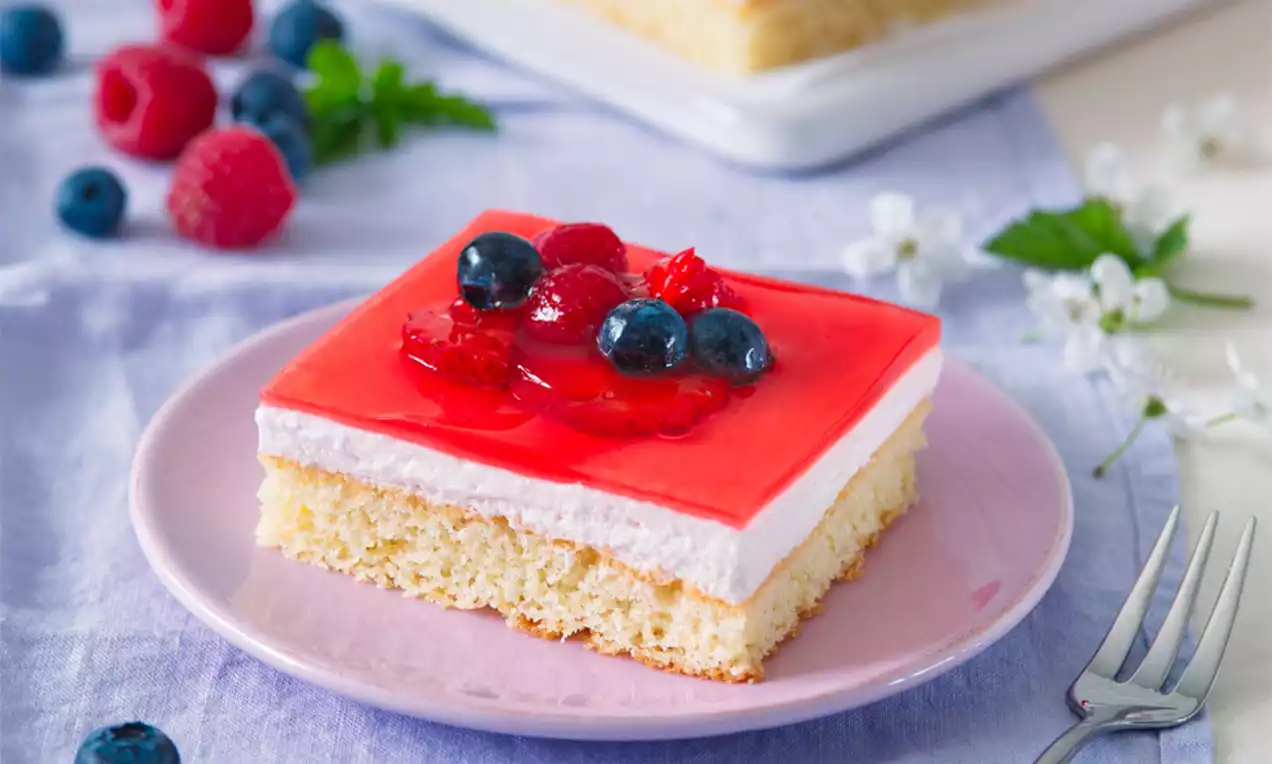 Čtvercový světlý zákusek s nadýchaným korpusem, vrstvou světlého krému a lesklým ovocným želé s borůvkami a malinami na růžovém talířku