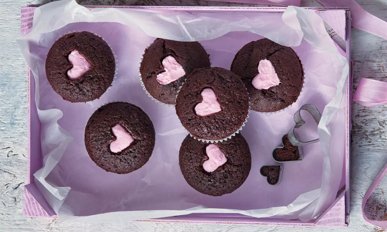 Čokoládové muffiny s matnou tmavohnedou textúrou, zdobené ružovými srdiečkami, naaranžované na fialovom papieri v rúžovom podnose