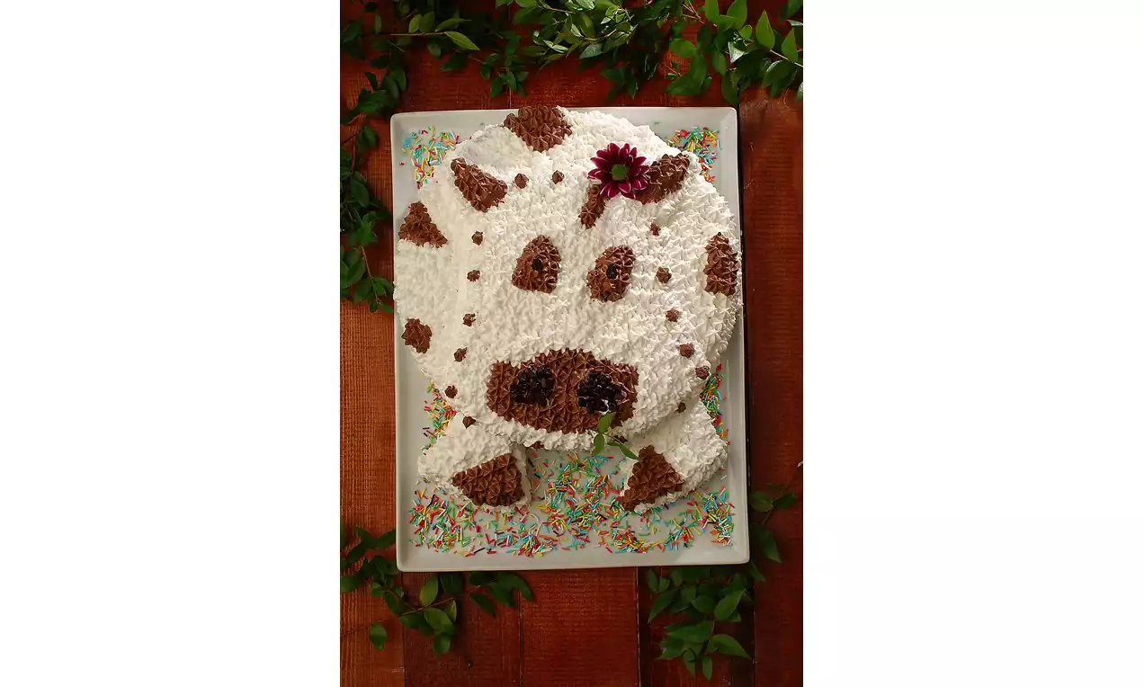 Beyaz kremalı ve kahverengi benekli, inek figürü şeklinde hazırlanmış pasta, renkli şekerlemelerle süslenmiş beyaz bir tabakta