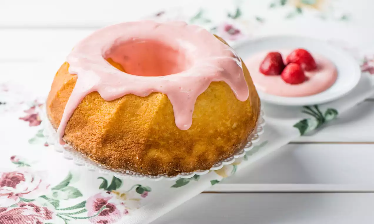 Jemne zlatistá bábovka s ružovým lesklým polevom, roztekajúcim sa po stranách, na čipkovanom podnose, vedľa tanierika s jahodami a omáčkou