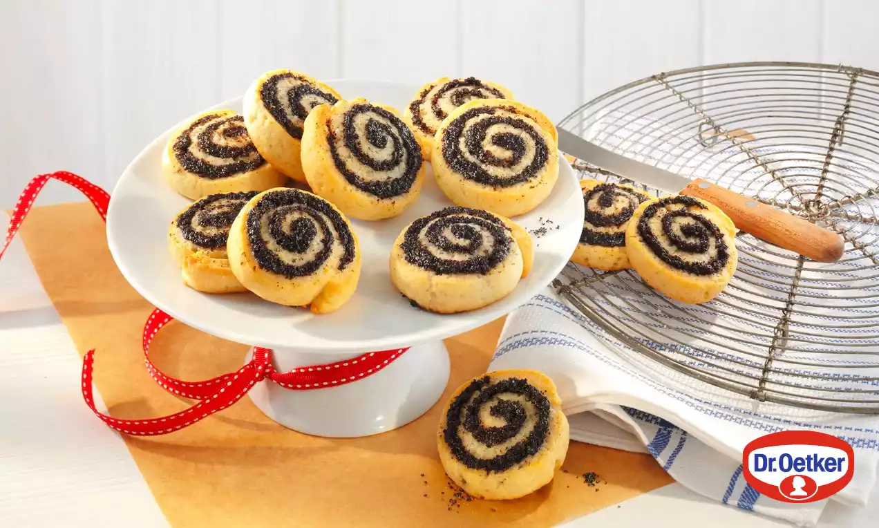 Makovo-pudingoví šneci s jemně zlatavou a měkkou texturou, spirálkový vzor černé náplně, naaranžovaní na bílém talíři vedle mřížky a červené stuhy