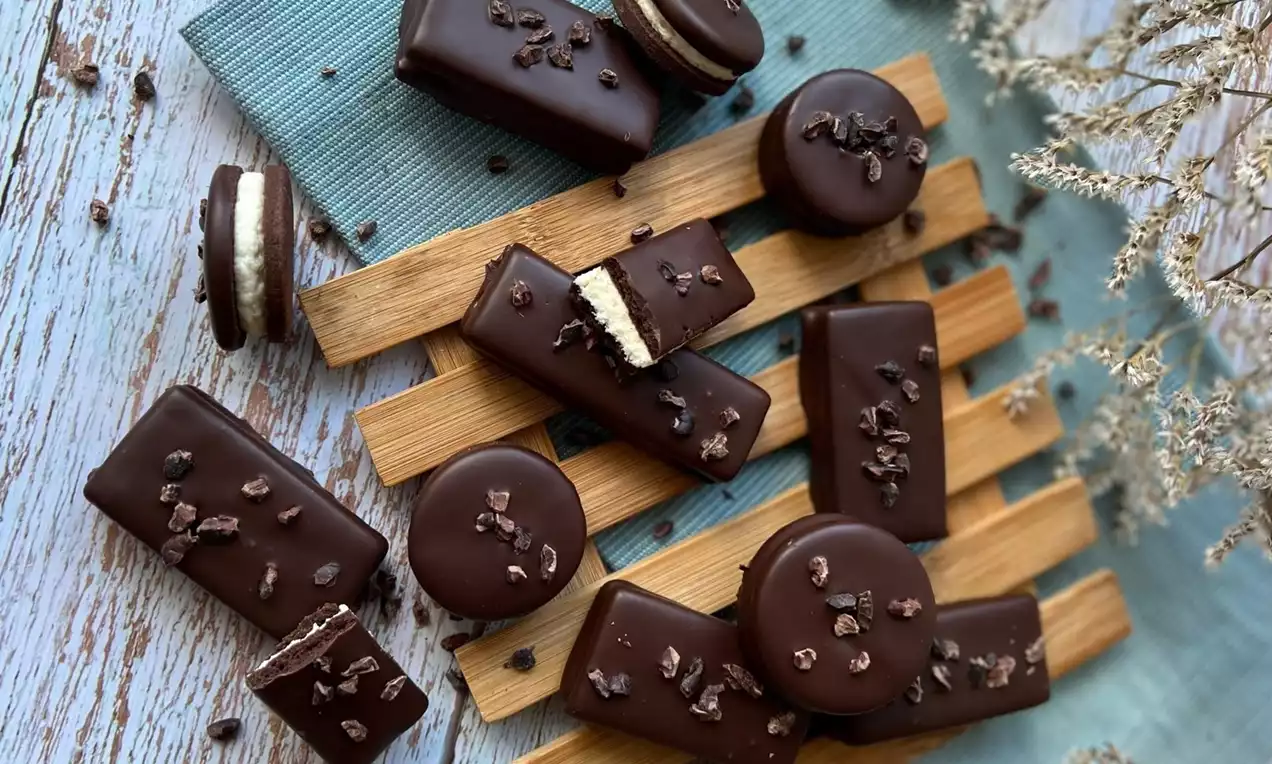 Lesklé marcipánové tyčinky v tmavé čokoládě, zasypané kakaovými kousky, uspořádané na dřevěné podložce vedle kulatých dezertů na látce