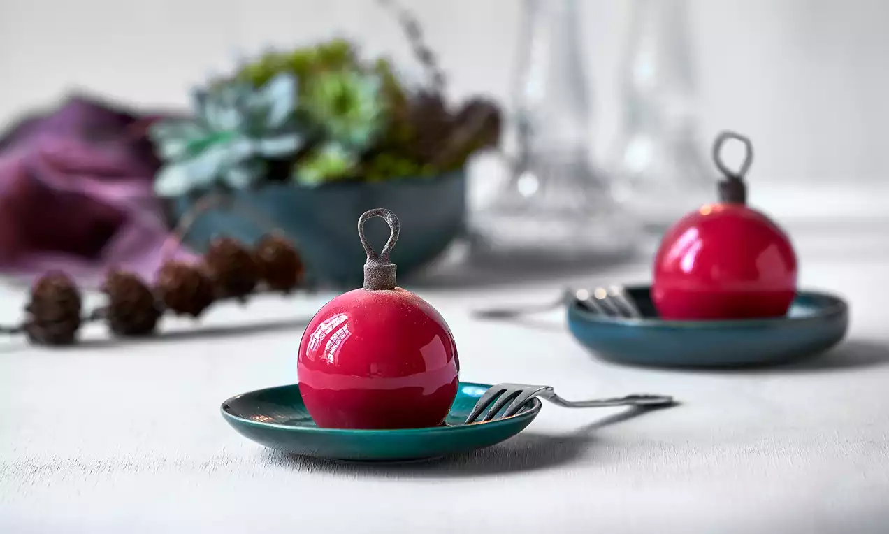 To glinsende røde julekugler med rund form og blank overflade på små mørkeblå tallerkener med gafler, omgivet af enkel borddekoration