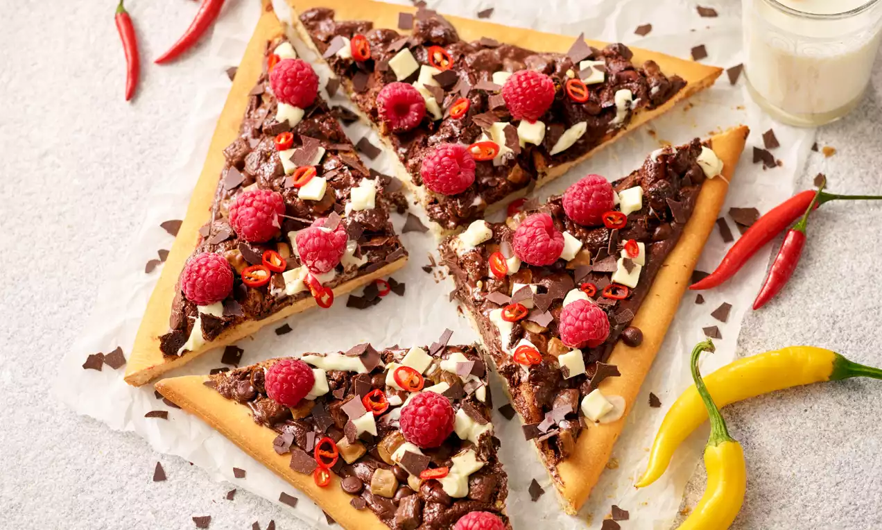 Knuspriger Pizzaboden mit geschmolzener Schokolade, Himbeeren, Chilischoten und Schokosplittern auf Backpapier angerichtet