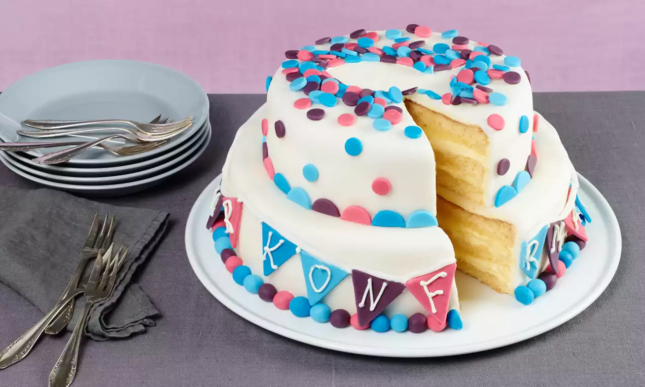 Zweistöckige Motivtorte mit weißem Fondant, bunten Zuckerperlen und Wimpeldekoration auf weißer Platte, ein Stück herausgeschnitten