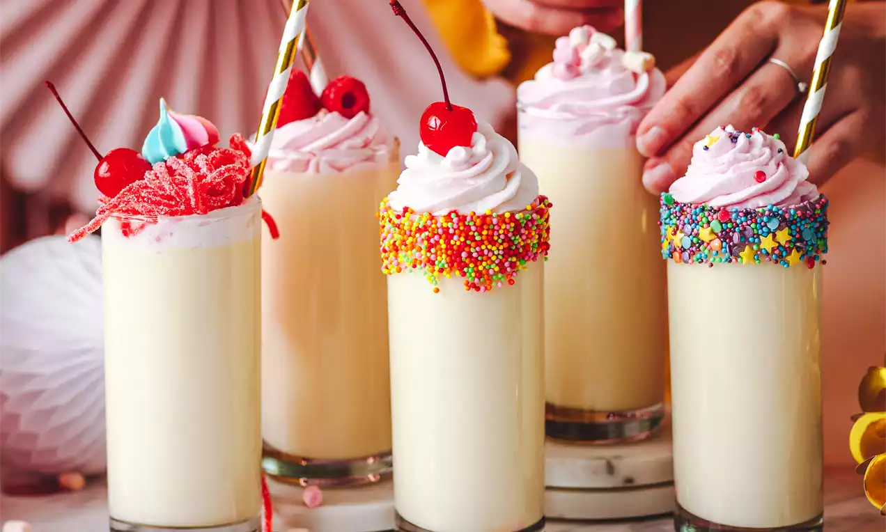 Vanilkový drink v průhledné sklenici s světlým krémovým mléčným základem, růžová šlehačka, zdobené posypy, třešně a papírová brčka