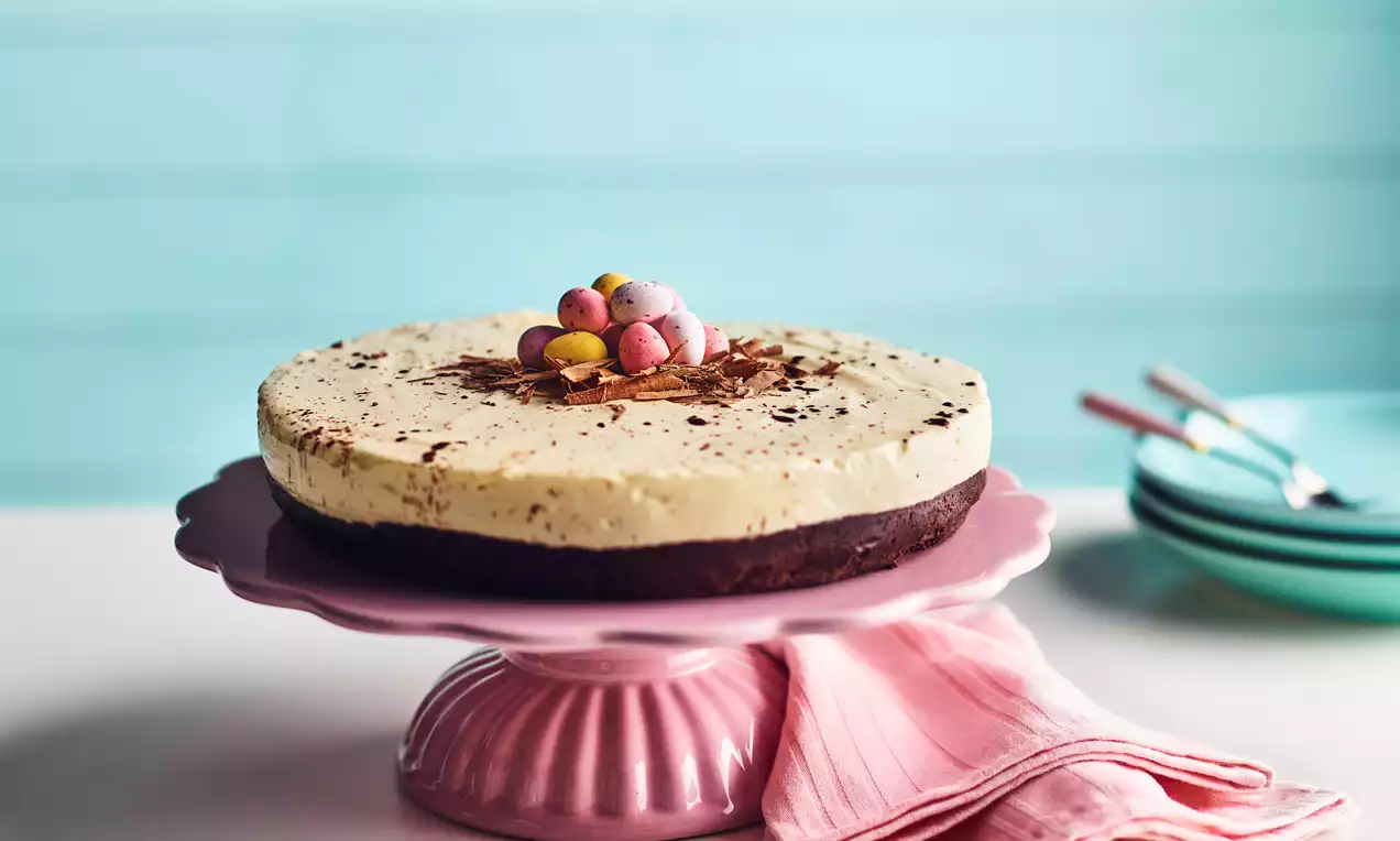 Round cheesecake with smooth pale cream surface, dark chocolate base, topped with colorful chocolate eggs and chocolate shavings on a pink stand