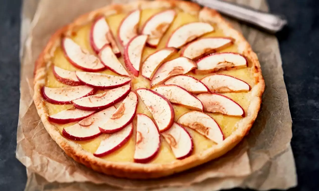Ohut ja pyöreä piirakka, jonka vaaleankeltaisella täytteellä lepää punareunaisten omenaviipaleiden symmetrinen kuvio ruskealla leivinpaperilla