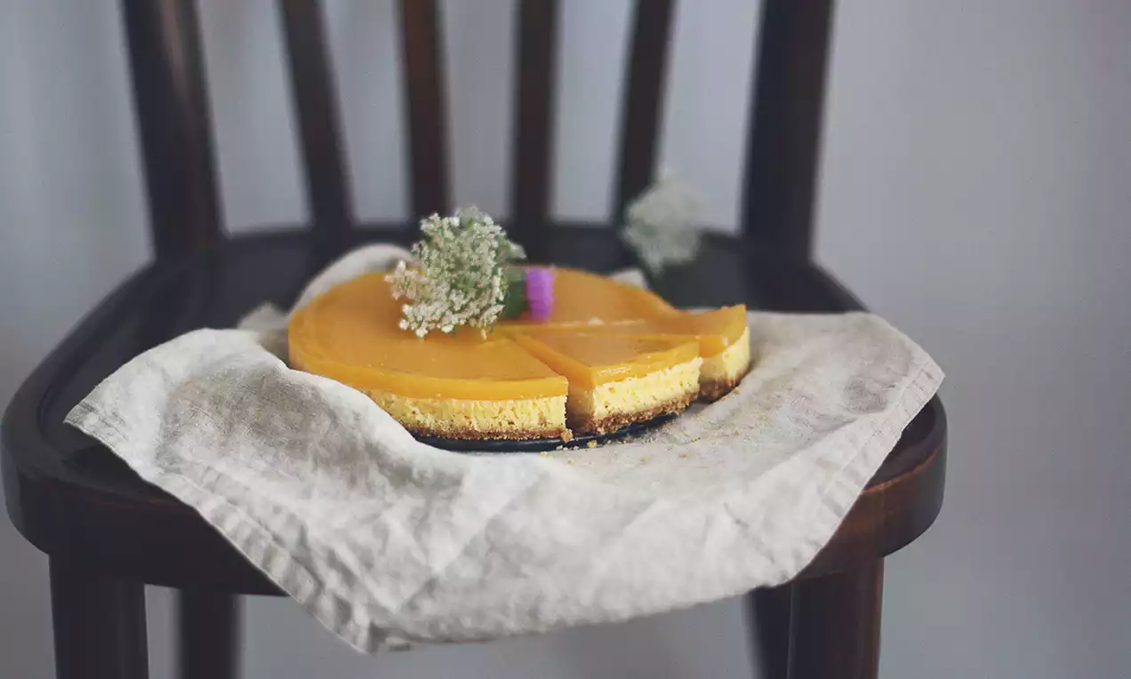 Kokosový cheesecake s hladkou žltou vrstvou manga na chrumkavom základe, ozdobený bielymi kvetmi, položený na ľanovej utierke na stoličke