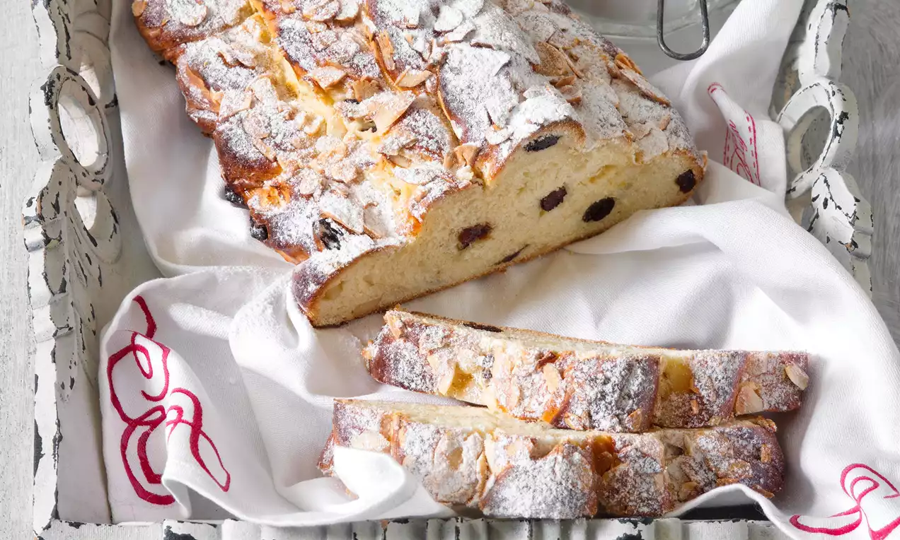 Bezvaječná vánočka s jemně zlatavou kůrkou, posypaná moučkovým cukrem, zdobená plátky mandlí, krájená na plátky v bílé textilní utěrce