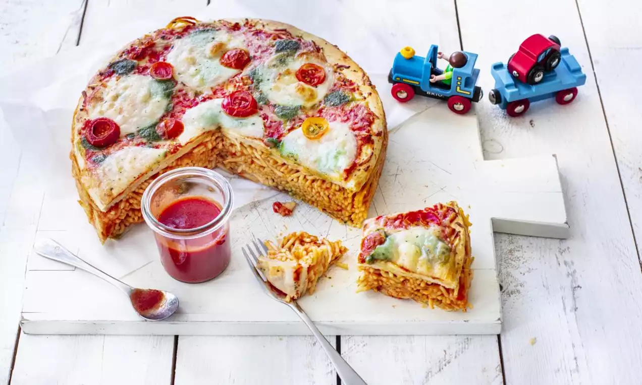 Round layered cake of spaghetti and tomato sauce topped with melted cheese, cherry tomatoes, herbs and served on a white board with a jar of tomato sauce beside