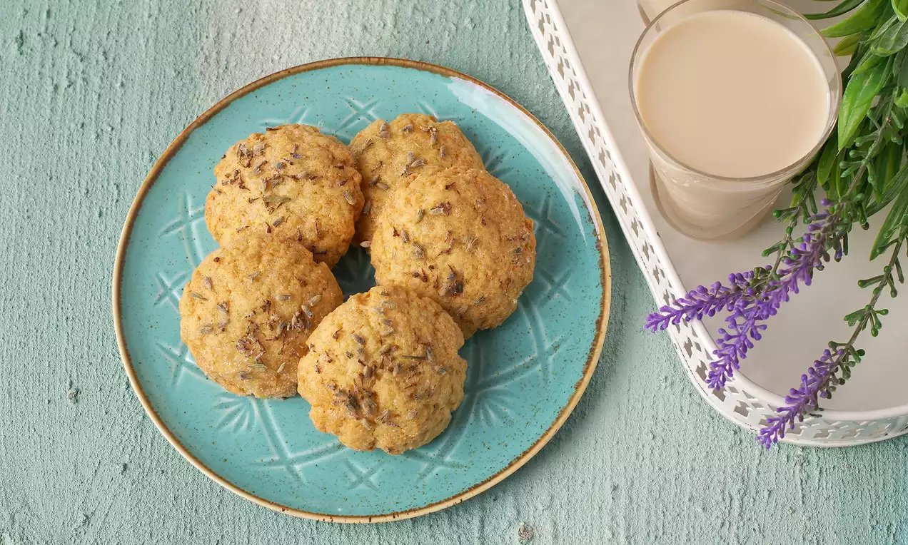 Lavanta taneleriyle süslenmiş altın rengi lavantalı kurabiyeler, turkuaz seramik tabakta düzenli şekilde yerleştirilmiş, yanında lavanta dalı ve süt