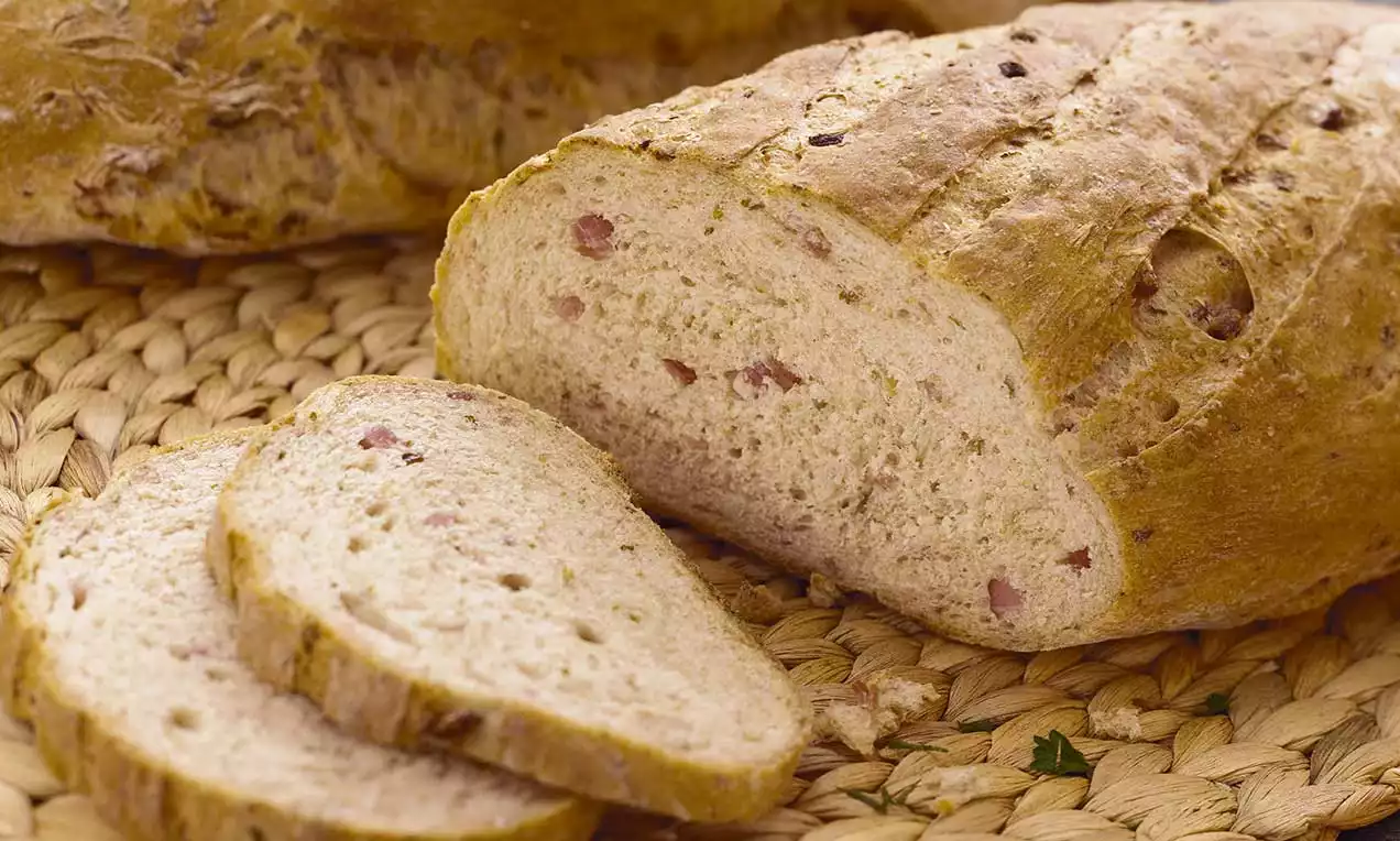 Pane rustico dorato con crosta croccante, interno morbido punteggiato di pezzi di speck rosa, affettato su un tappetino di paglia intrecciata