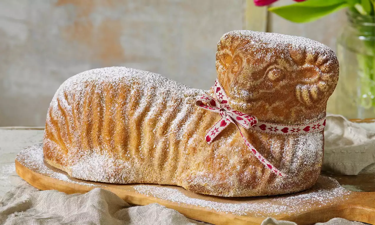 Citronový velikonoční beránek zlatavé barvy s jemně zrnitou texturou, posypaný moučkovým cukrem, ozdobený mašlí na dřevěném podnosu