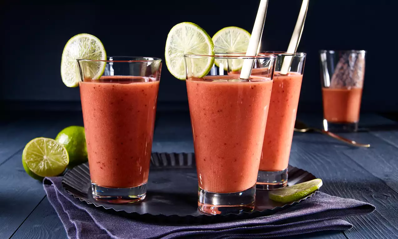 Fruchtiger Smoothie in kräftigem Rot, serviert in klaren Gläsern mit Limettenscheiben und Strohhalmen, arrangiert auf blauem Tuch