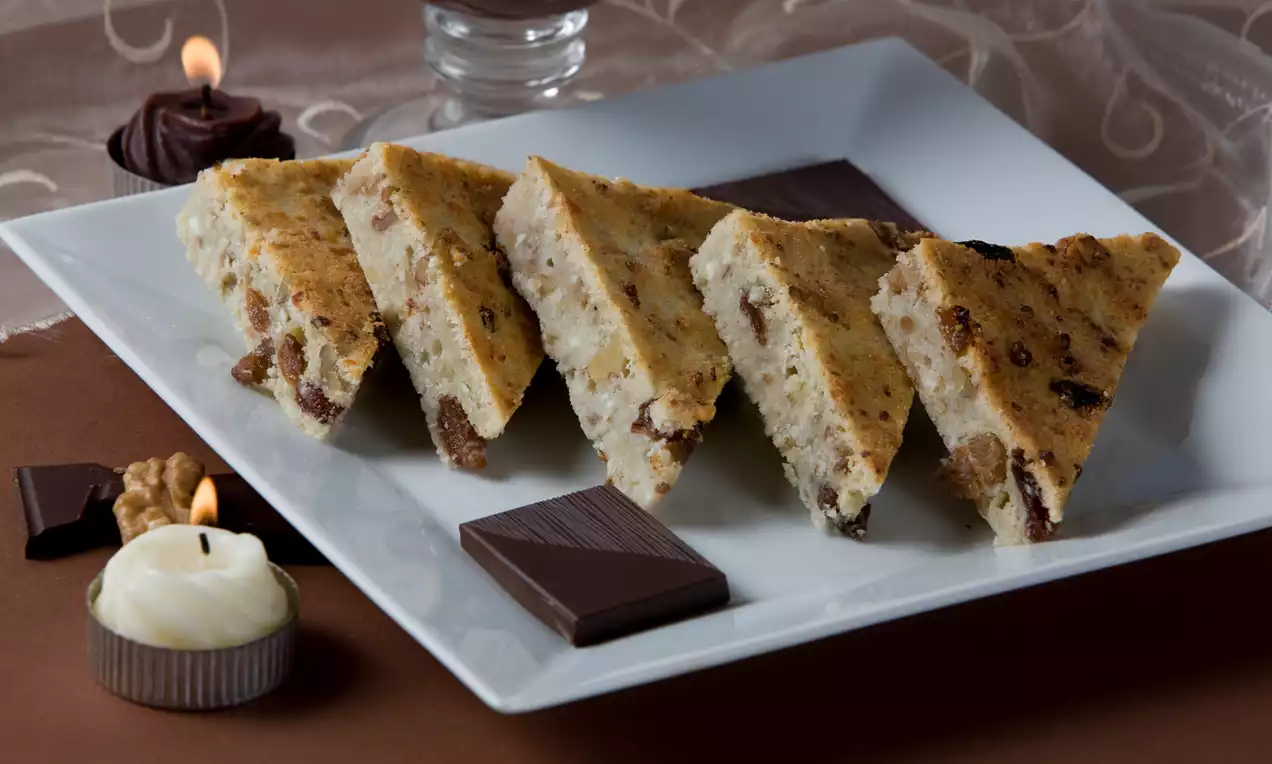 Trojuhoľníkové kúsky tvarohového chlebíčka svetlej farby s hnedými kúskami hrozienok, položené na bielom tanieri vedľa čokolády a sviečok