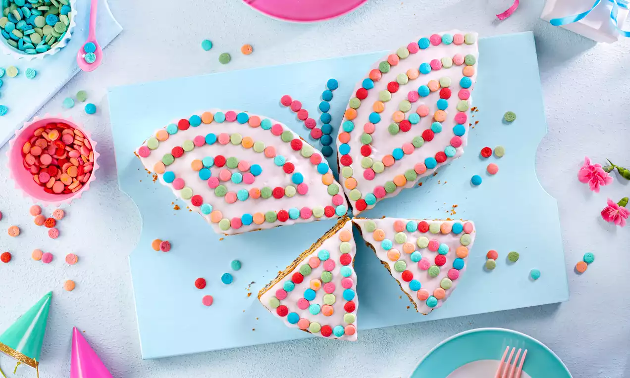 Schmetterlingskuchen mit heller Glasur, verziert mit bunten Schokolinsen auf einer blauen Platte, umgeben von farbenfroher Partydekoration