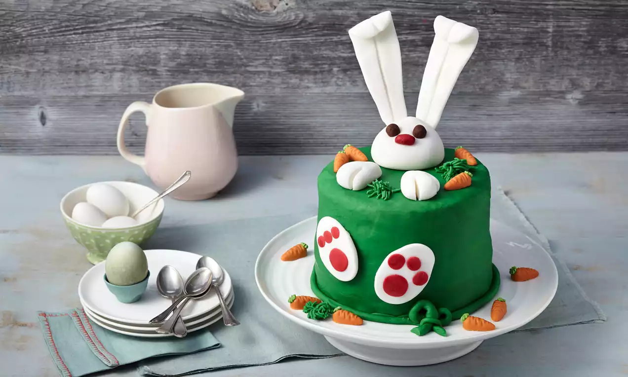 Petite tourte verte recouverte de pâte à sucre avec un lapin blanc en relief, des carottes miniatures et des décorations colorées sur une assiette blanche