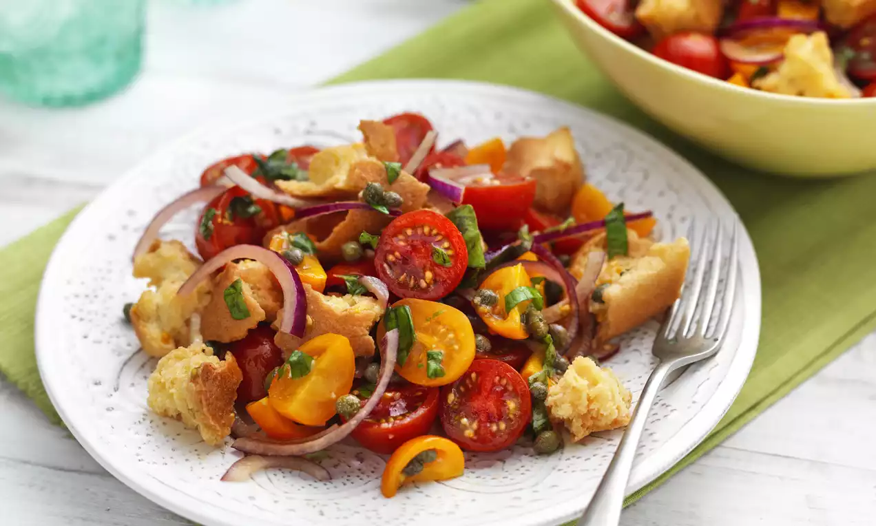 Bright panzanella with red and yellow tomato slices, crisp bread chunks, purple onion, and basil leaves on a white patterned plate