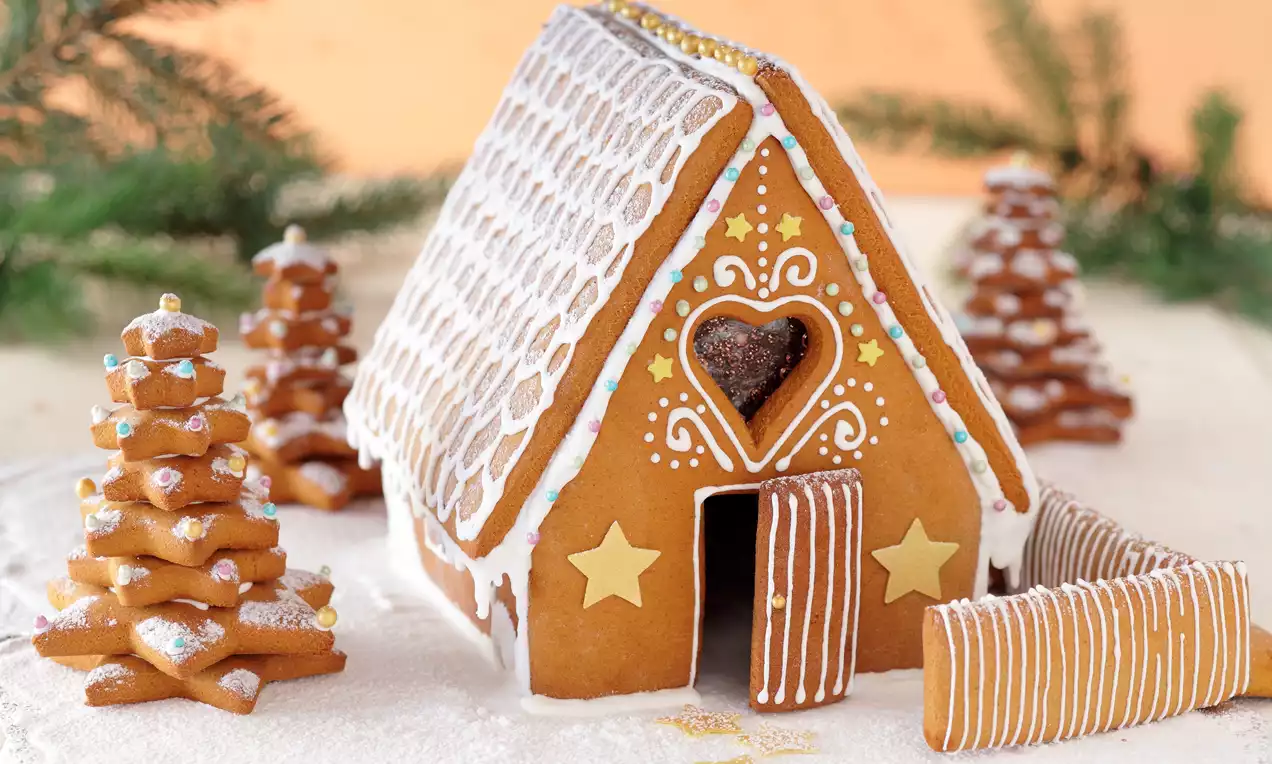 Piernikowa chatka z białym lukrem, złotymi gwiazdkami i sercem na fasadzie, otoczona piernikowymi choinkami na śnieżnym tle z gałązkami jodły
