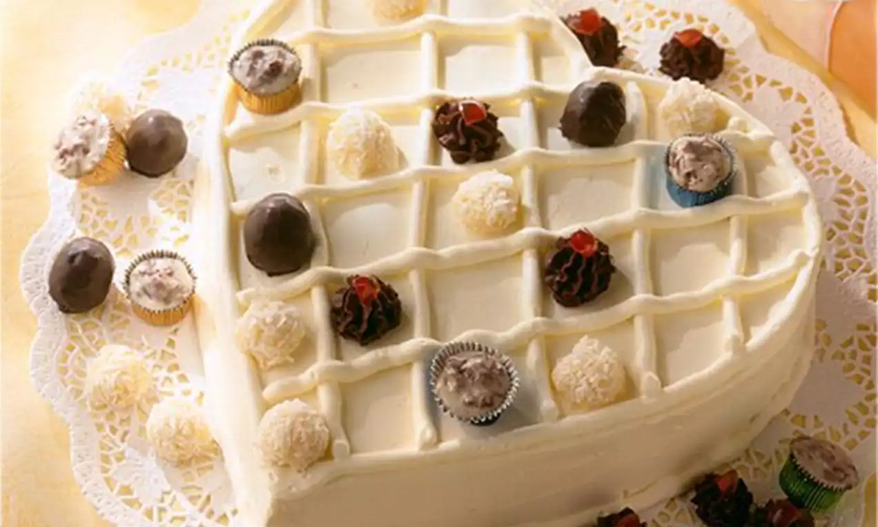 Gâteau en forme de cœur couvert d’un glaçage blanc lisse avec un motif en quadrillage, décoré de truffes et de rochers chocolatés