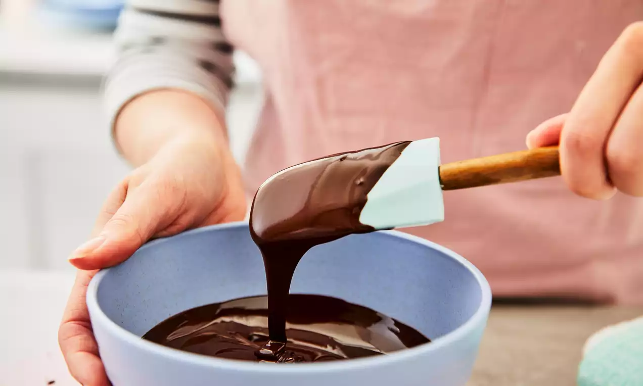 Glossy melted chocolate in rich dark brown flowing smoothly from a spatula into a pale blue bowl held with both hands