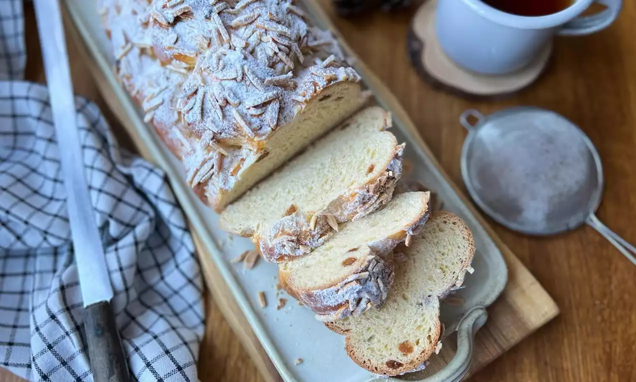 Vánočka jemně zlatavé barvy, posypaná moučkovým cukrem a plátky mandlí, nakrájená na silné krajíce na podlouhlém talíři