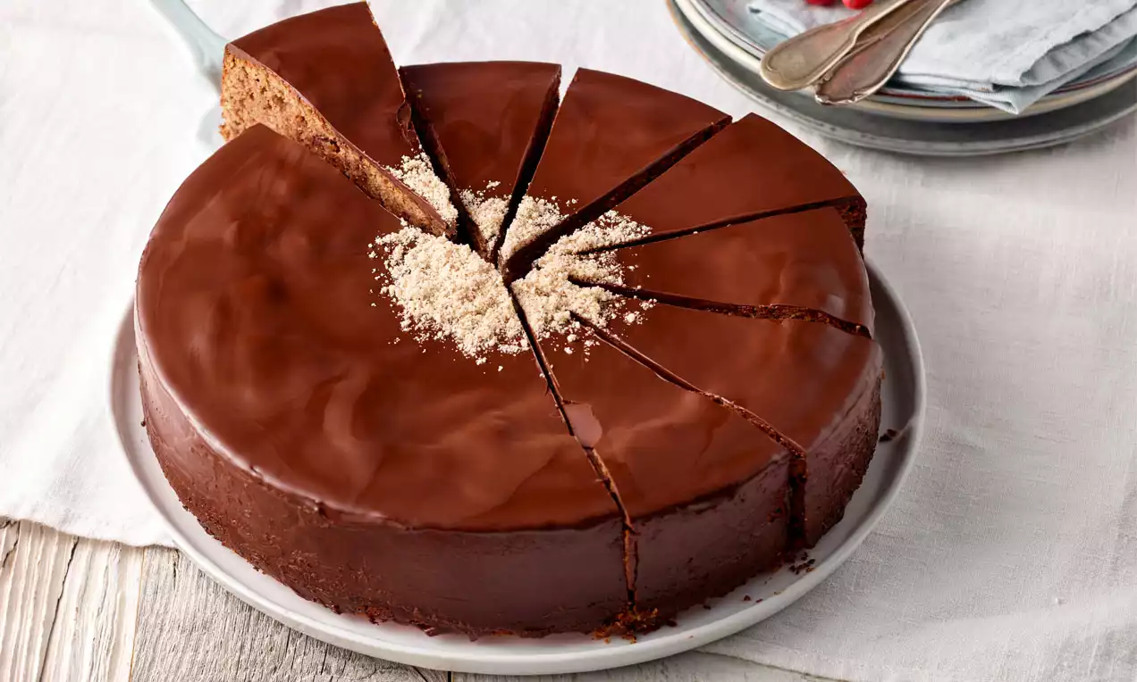Runder Maroni-Mandel-Kuchen mit glänzender Schokoladenglasur, in Stücke geschnitten, dekoriert mit gehackten Mandeln auf weißem Teller