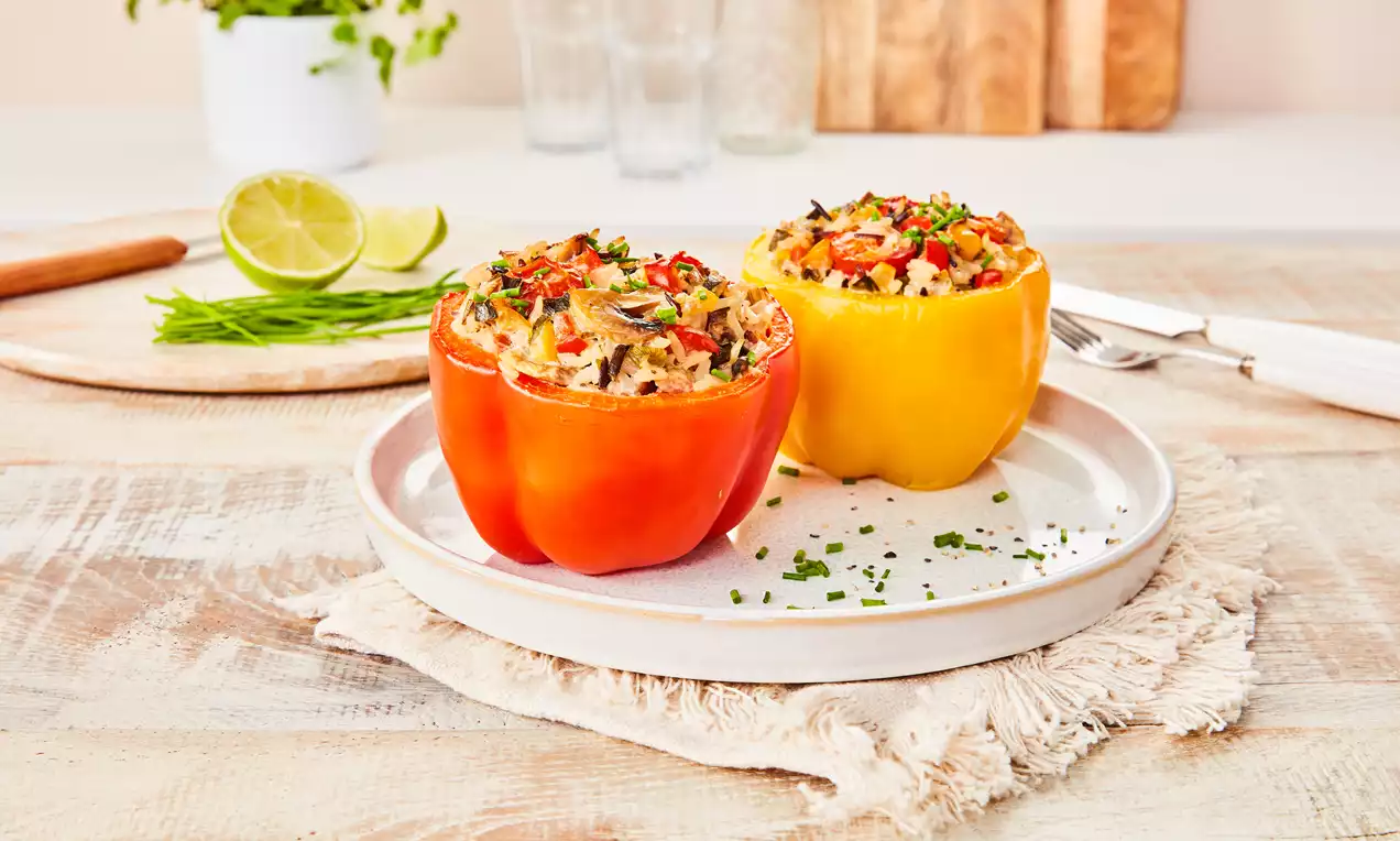 Zwei gefüllte Paprika in Rot und Gelb, auf weißem Teller angerichtet, mit Reisfüllung und Kräutern garniert, auf heller Holzoberfläche