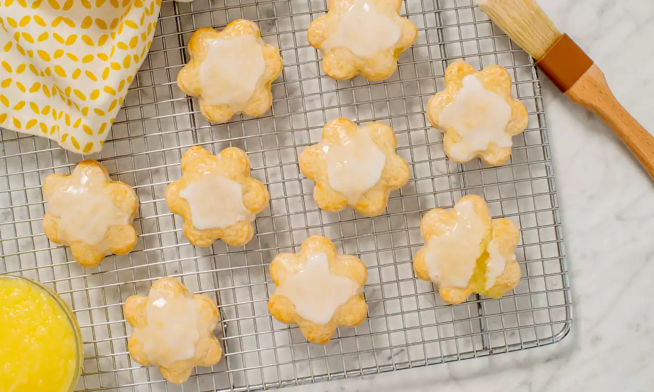 Flower-shaped lemon cookies with golden edges and glossy white icing arranged on a wire rack, beside a bowl of lemon curd and a pastry brush