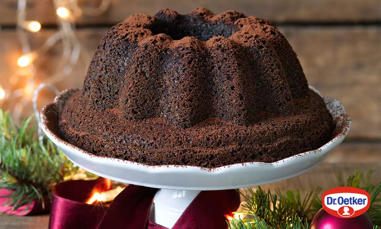 Bezgluténová maková bábovka tmavohnedej farby s výraznou vrúbkovanou textúrou na bielom tanieri, ozdobená červenou stuhou a vetvičkami.