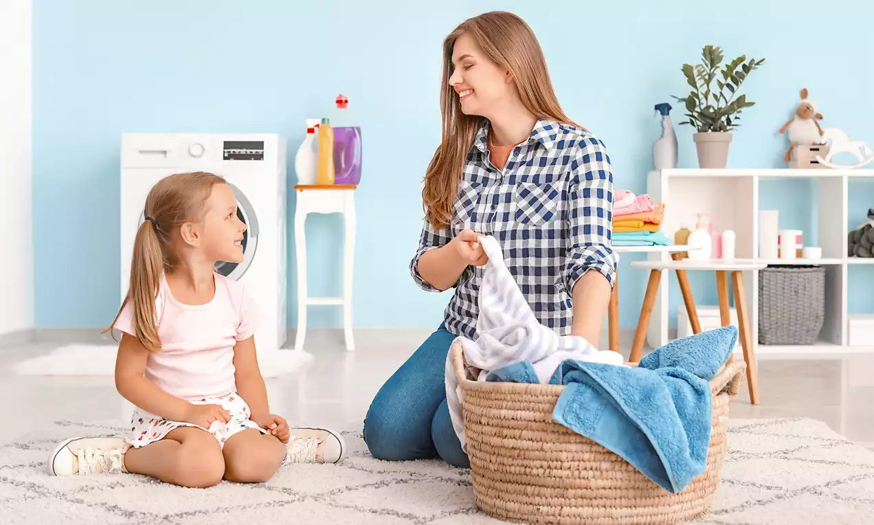 A woven basket filled with soft white and blue cloths sits on a textured gray carpet in a bright room with pastel blue walls and tidy shelves