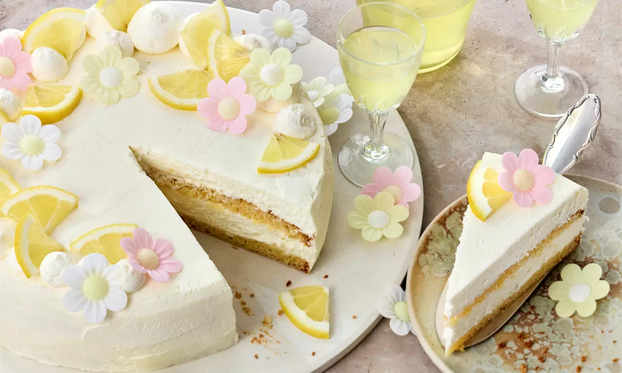 Helle Limoncello-Torte mit cremiger Schichtfüllung, dekoriert mit Zitronenscheiben und pastellfarbenen Zuckergänseblümchen auf weißer Platte