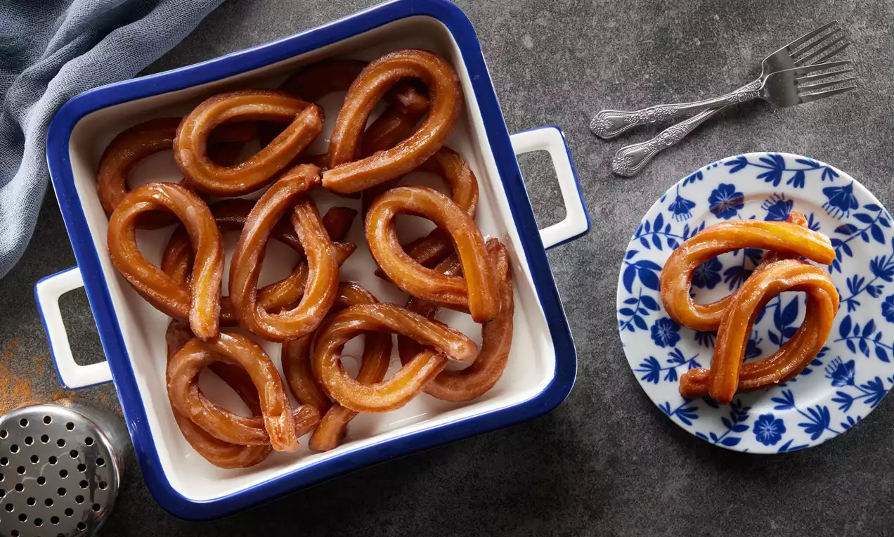 Altın rengi şerbetle kaplanmış halka tatlıları beyaz kenarlı kare bir tepsi ve desenli mavi beyaz tabakta sunulmuş