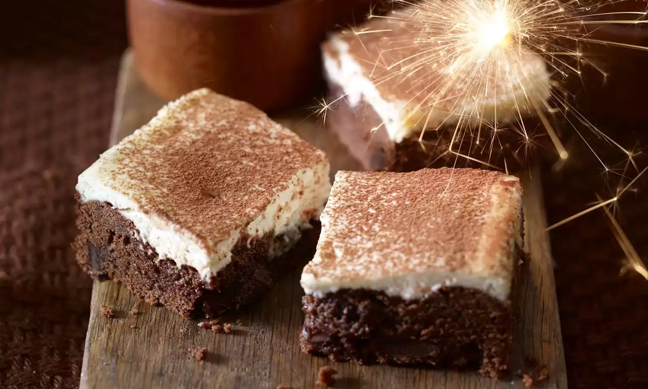 Carrés de brownie chocolat moelleux recouverts d’une couche de crème blanche saupoudrée de cacao sur une planche en bois