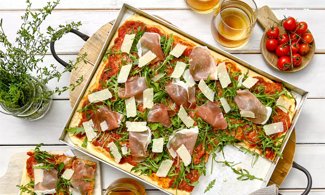 Pravougaona pizza s tankom korom, crvenim umakom, rikulom, tankim kriškama pršuta i svijetlim komadima sira na drvenoj podlozi