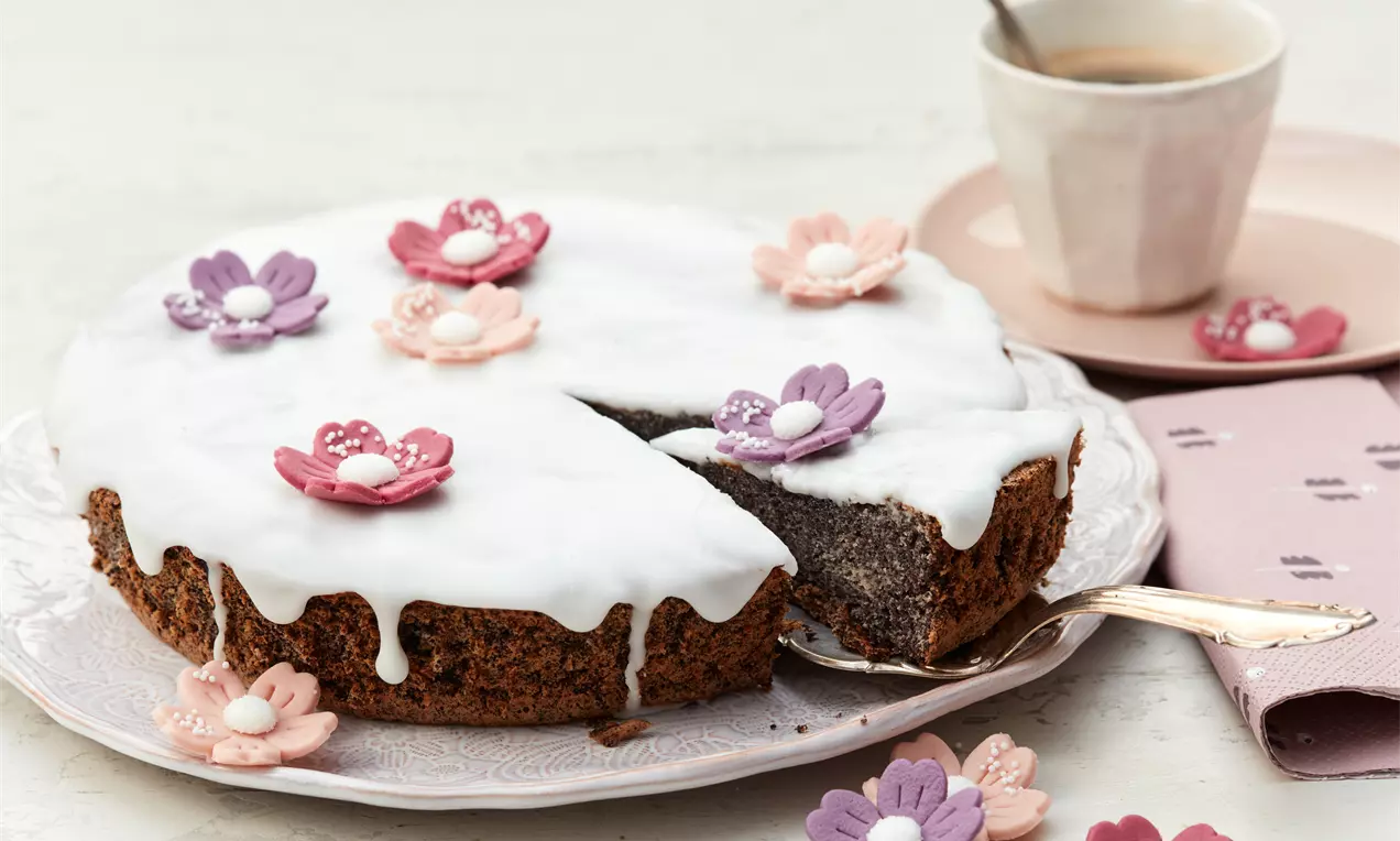 Runder Mohnkuchen mit weißem Zuckerguss und bunten Blüten aus Zuckerdekor auf einem Teller, daneben rosa Serviette und Tasse Kaffee