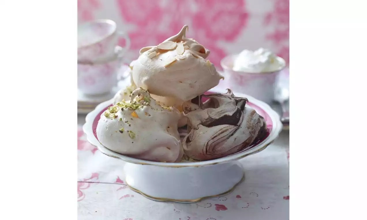 Swirled mini meringues in shades of cream, brown, and ivory, topped with pistachio pieces, stacked on a decorative pedestal plate