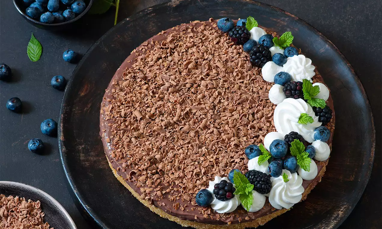 Kulatá pie s hladkou čokoládovou polevou, ozdobená strouhanou čokoládou, šlehačkou, čerstvými borůvkami, ostružinami a lístky máty na tmavém talíři