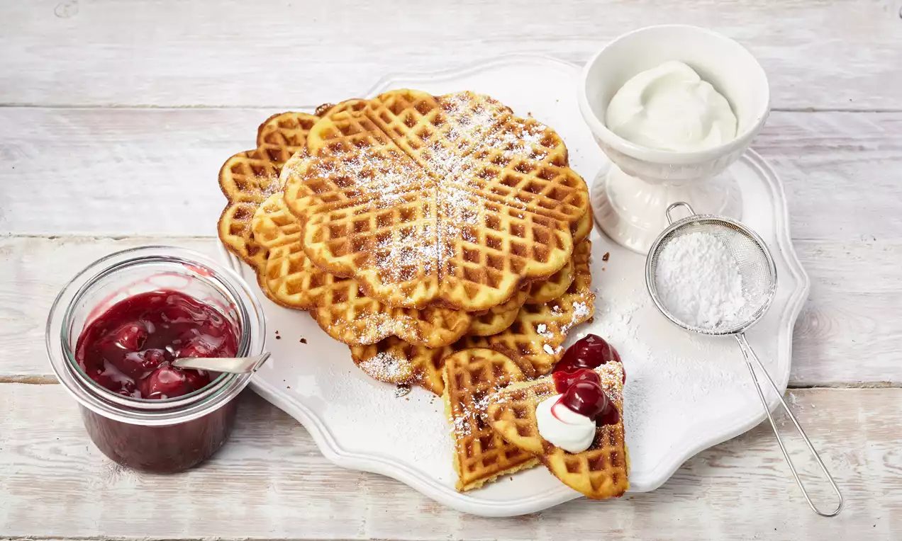 Goldgelbe, herzförmige Waffeln auf weißer Platte mit Puderzucker bestäubt, serviert mit Kirschkompott und Sahne in Schale
