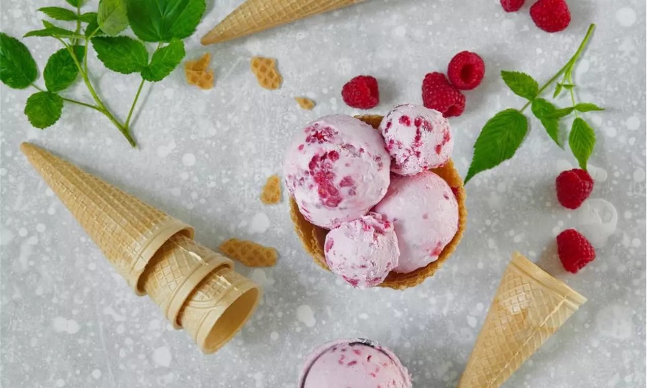 Malinová smotanová zmrzlina jemne ružovej farby s kúskami malín, servírovaná v kornútoch a miske, obklopená čerstvými malinami a listami mäty