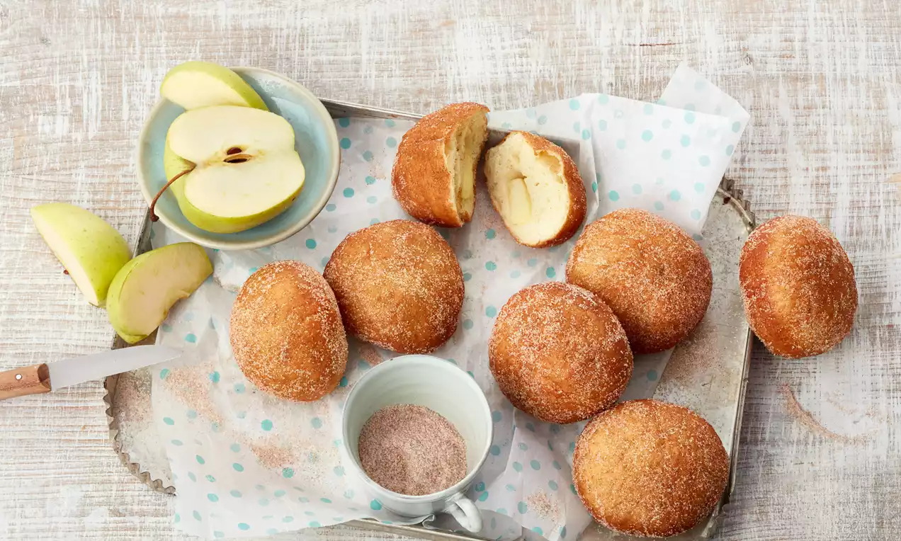 Apfel-Krapfen, goldbraun in Zimt-Zucker, ein aufgeschnittener Krapfen zeigt saftige Apfelstücke und Puderzucker auf hellem Teller