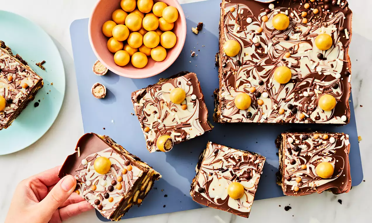 Billionaire Tiffin squares with marbled milk and white chocolate tops, golden spheres, and crunchy biscuit layers on a blue board